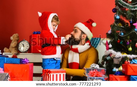 Christmas family opens presents on red background. Man with beard and busy face plays with son. Winter holiday and celebration concept. Santa and little assistant among gift boxes near Christmas tree