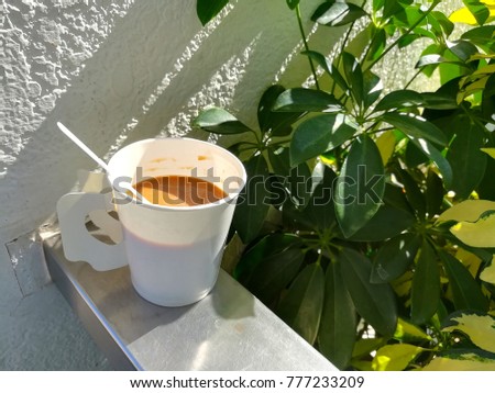 A white coffee cup rests on a railing with yellowish green leaves as the background