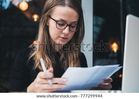 Portrait of a young beautiful girl sitting at table with laptop, holding pen in hand  and reading piece of paper. Education concept