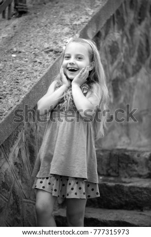 small baby girl or cute happy child with adorable smiling surprised face and bow in blonde hair in blue dress in summer outdoor near stony stairs on blurred background