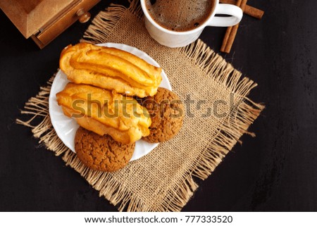 A cup of coffee, eclairs and oatmeal cookies on a black textured background.