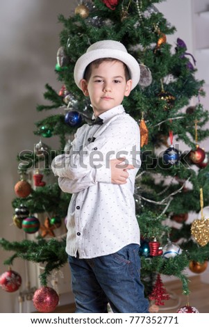 boy with serious attitude with cross arms in a blue jeans and white shirt with black bow tie and festive white hat celebrating Christmas standing alongside a decorated Christmas tree 