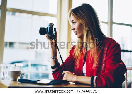 Professional female photographer taking picture on old camera focusing lens concentrated on work.Hipster girl fond of photography using vintage equipment making images out of window sitting in cafe