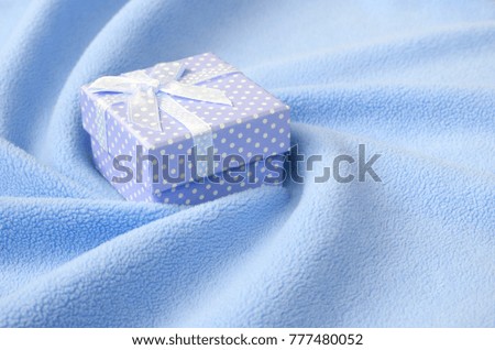 A small gift box in blue with a small bow lies on a blanket of soft and furry light blue fleece fabric with a lot of relief folds. Packing for a gift to your lovely girlfriend
