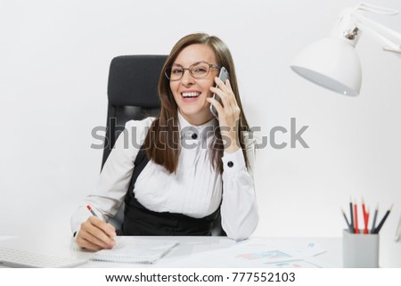 Smiling business woman in suit sitting at the desk, working at contemporary computer with document in light office, talking on mobile phone, conducting pleasant conversation on white background