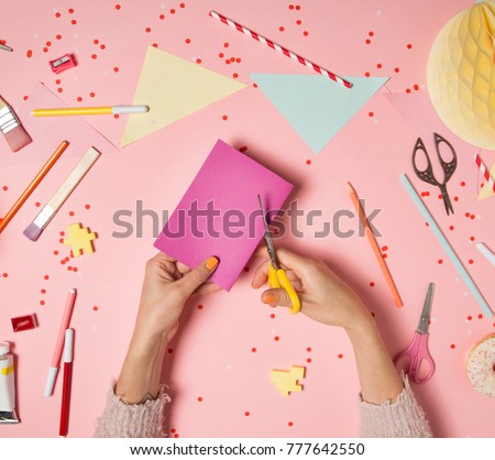 Colorful pink background with various party confetti, paper decoration, flags, stationary, DIY accessories with woman's hands cutting paper card. Handmade concept flat lay top view. Party arrangement Royalty-Free Stock Photo #777642550