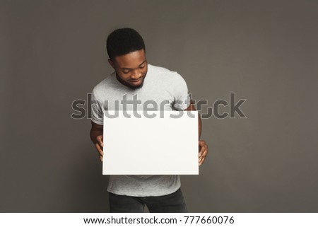 Picture of young smiling african-american man holding white blank board on grey background, copy space