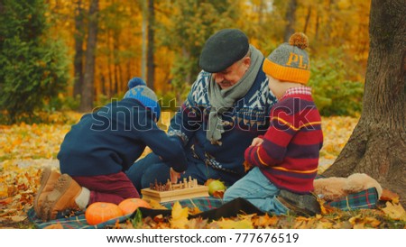 Little boys are playing chess with his grandfather in the autumn park under the big tree