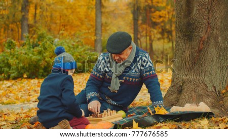 Little boy is playing chess with his grandfather in the autumn park under the big tree