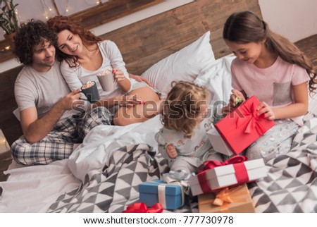 It's Christmas time! Happy family sitting in bed, parents are drinking hot cocoa while their daughters are opening the presents