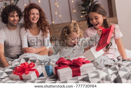 It's Christmas time! Happy family sitting in bed, parents are smiling while their daughters are opening the presents
