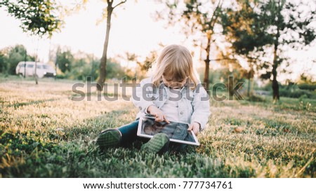 A cute picture of small little girl sitting on grass and playing with big tablet. She wants to watch some cartoons on it so that's why she is pointing on the screen.