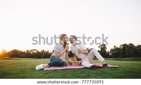 Another picture of two young and fallen in love people sitting on mat in green and beautiful park. He is smiling and looking at her at the same time while she is drinking some juice and looking at him