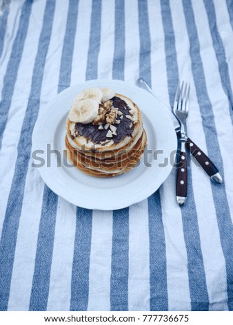 Delicious homemade pancakes with chocolate nougat, sliced banana, nuts. Striped cloth, fork and knife, wooden background. Breakfast outside, picnic, morning nutrition. Space for text.