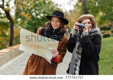 Portrait of two attractive girls dressed in autumn clothes holding city map while standing outdoors and making photos with vintage camera