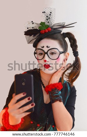 A young pretty girl mime, in a lady's hat and bright suit, makes selfie on her smartphone, grimacing, she parodies narcissism in women nowadays