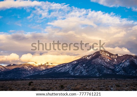 Amazing sunset view of Taos , New Mexico Mountains outside of the city as clouds roll in and the sun paints a golden picture as it sets with snow along the grand and winter shrubs