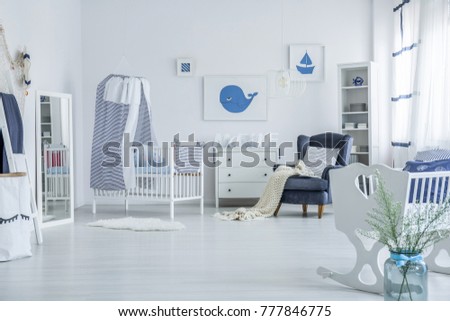 Vase next to white cradle in spacious baby's room with blanket on armchair and striped veil above crib