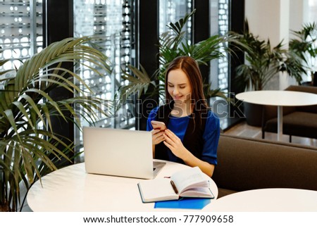 young nice business woman works behind the laptop for little tables in cafe and talks on the smartphone. Nearby there are a cup of coffee and the daily log with records.