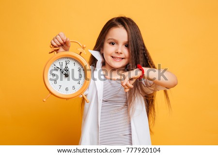 Image of cheerful little girl child standing isolated over yellow background. Looking camera pointing to clock alarm.