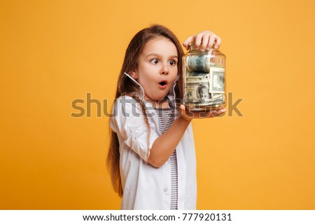Photo of shocked little girl child standing isolated over yellow background holding jar with money. Looking aside.
