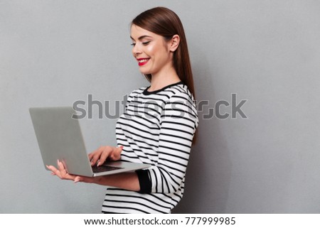 Portrait of a happy woman using laptop computer while standing isolated over gray background