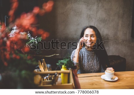 beautiful young brunette girl talking on mobile phone at wooden table near window and drinking coffee in cafe decorated with Christmas decor. Dressed in a gray knitted wool sweater.