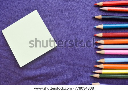 Multicolored, bright, colorful pencils for drawing located on the right and a notebook on the left for your text on a background of purple cloth.