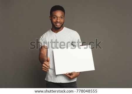 Picture of young smiling african-american man holding white blank board and showing thumb up on grey background, copy space