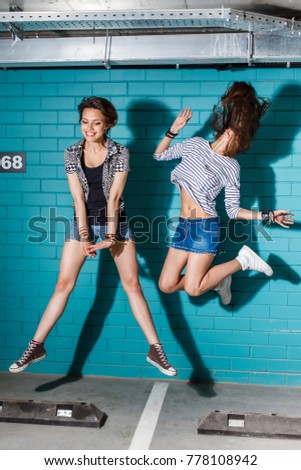 Lifestyle portrait of two beautiful best friend hipster girls wearing stylish bright outfits and denim shorts. Jumping together at parking in front of blue brick wall and have great time and fun.