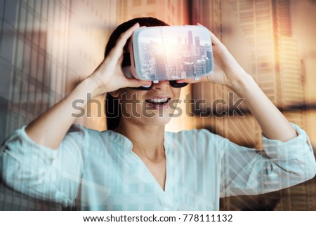 New experience. Upbeat young woman in a white blouse wearing a virtual reality headset and touching it with both hands while smiling