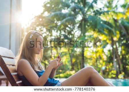 Happy smartphone woman relaxing near swimming pool listening with earbuds to streaming music. Beautiful girl using her mobile phone app 4g data to play songs while relaxing on summer luxury vacations.