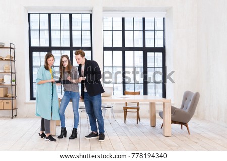 Creative teamwork. Positive office workers - two women and man stand near large wooden table and look at the tablet discussing new project on the background of large windows in office