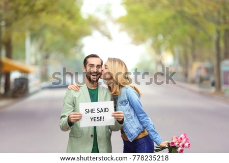 Young man holding paper with text SHE SAID YES and his happy beloved on engagement day outdoors Royalty-Free Stock Photo #778235218