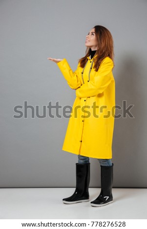 Full length portrait of a smiling girl dressed in raincoat and rubber boots standing with outsretched hand isolated over gray background