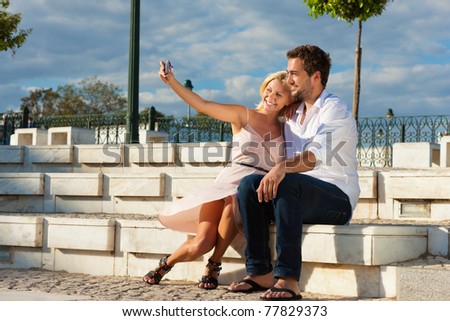 Couple having a city break in summer sitting on a brick wall in the sunlight, making pictures