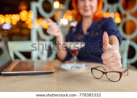 A woman, sits in a cafe and shows a gesture of thumbs up.