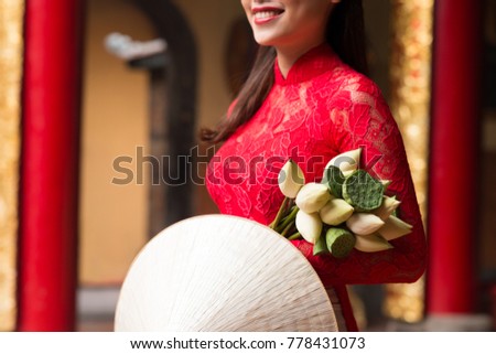 Close-up shot of unrecognizable woman wearing red lace dress looking away with toothy smile while holding conical hat and flower bouquet in hands, interior of temple on background