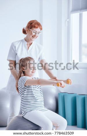 Smiling girl exercising with orange dumbbells with therapist's support