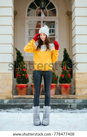 Full heigh portrait of inspired woman in warm yellow sweater and red mittens walking on the european city. Outdoor photo of hipster brunette woman in white knitted hat spending time outdoor.