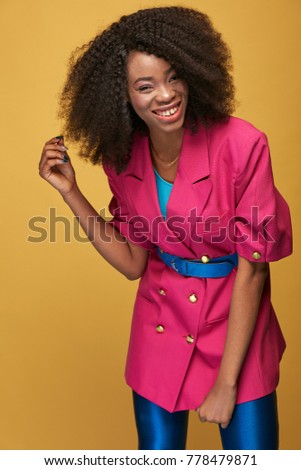 Beauty portrait of attractive young african girl with afro hairstyle. Smiling girl wearing pink jacket, blue leggings and belt posing on yellow background. Studio shot