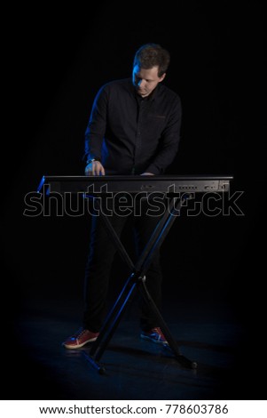 Young blond man in black clothes playing on a musical synthesizer and posing on a black background with blue light