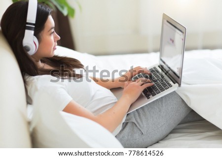 Young pretty woman has a rest on the bed with laptop and listing a music