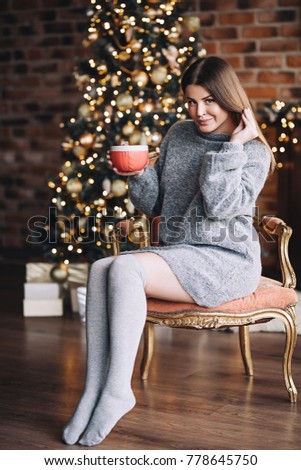 Happy smiling woman in the sweater drinks hot chocolate near the shining christmas tree at her living room, christmas eve, home comfort and New Year's mood, warming drink, pretty attractive woman