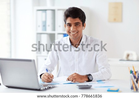 business and people concept - businessman in earphones with papers and laptop computer working at office and listening to music