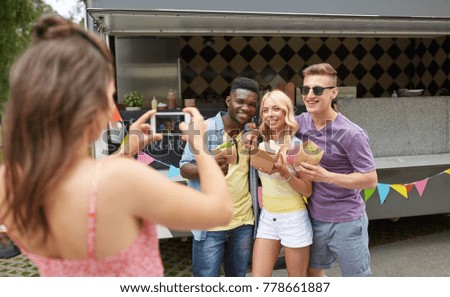 leisure, technology and people concept - young woman taking picture of her happy friends eating hamburgers and wok at food truck
