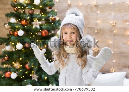 Beautiful blonde girl, has happy fun smiling face, grey hat pullover, Christmas tree lights. Portrait holiday. Winter background. Family kids portrait. New year decoration. 