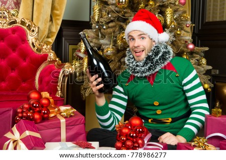 Man in santa hat smile with champagne bottle. Gift giving, boxing day. Happy new year, xmas, holidays. Celebration, party concept. Macho in elf costume with present boxes at Christmas tree.