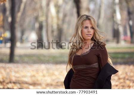 Outdoor portrait of a beautiful long-haired blond young woman wearing a black coat, posing in a park in late fall. Shallow depth of field.