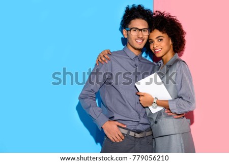 Young african amercian businesswoman holding tablet and hugging stylish businessman on pink and blue background 